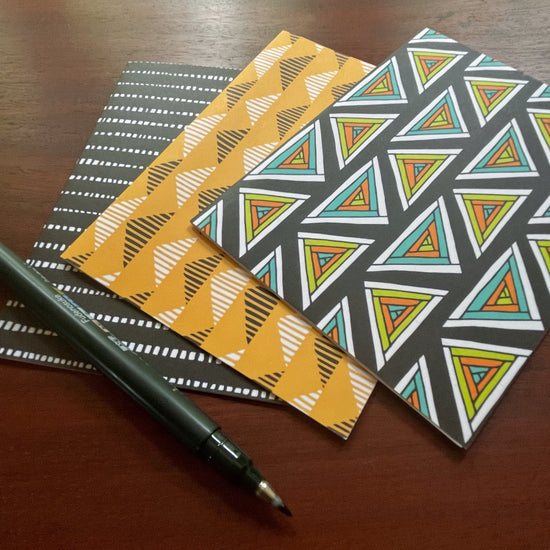 geometric patterned notecards fanned out with pen. Bottom card is black with white stripes of irregular squares. Middle card is yellow with rows of black and white striped triangles. Top card is black with rows of colorful concentric triangles