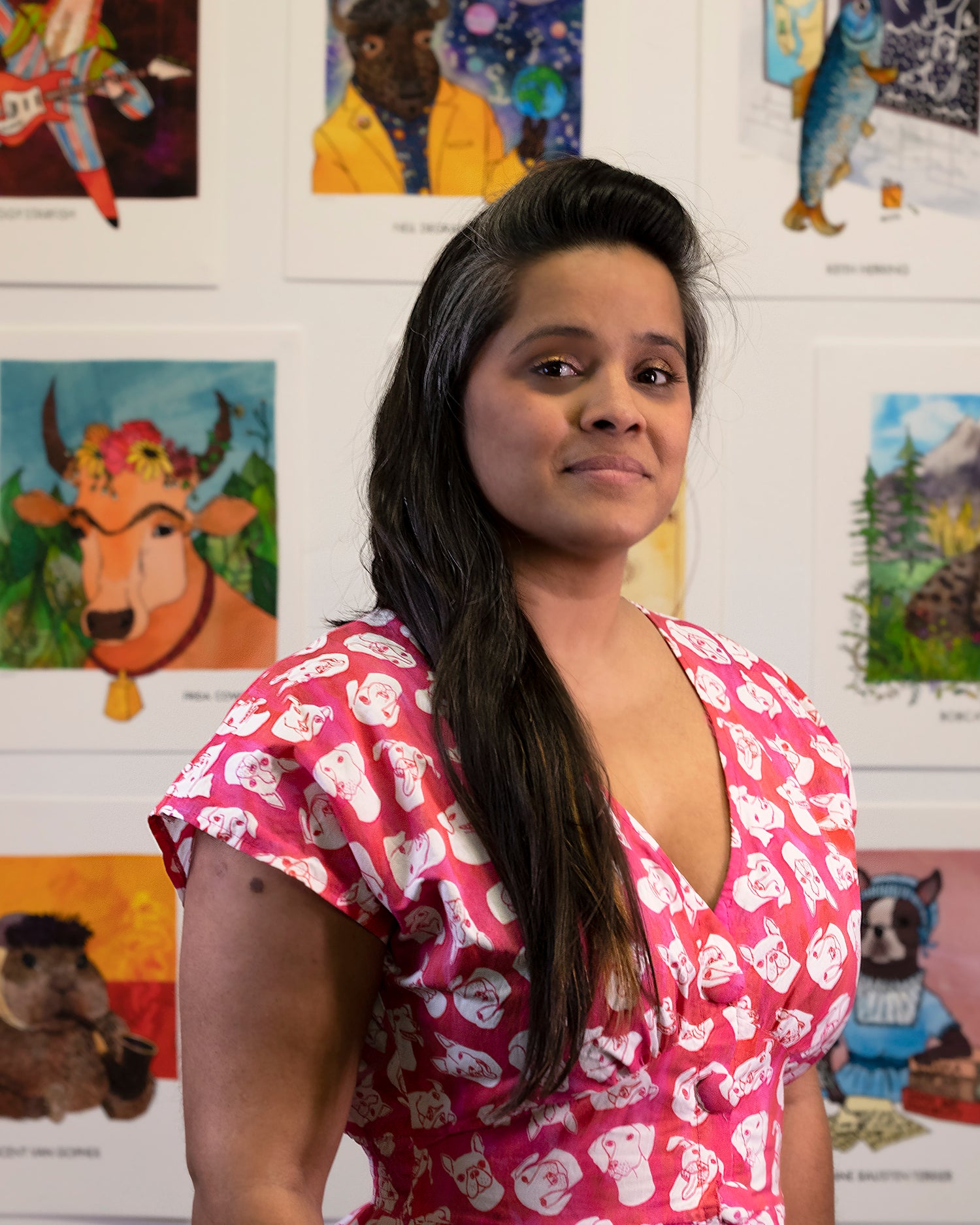 headshot of Krishna Chavda in pink dress with white dog heads. she's standing in front of a colorful background of her art prints featuring the Famous Animals series