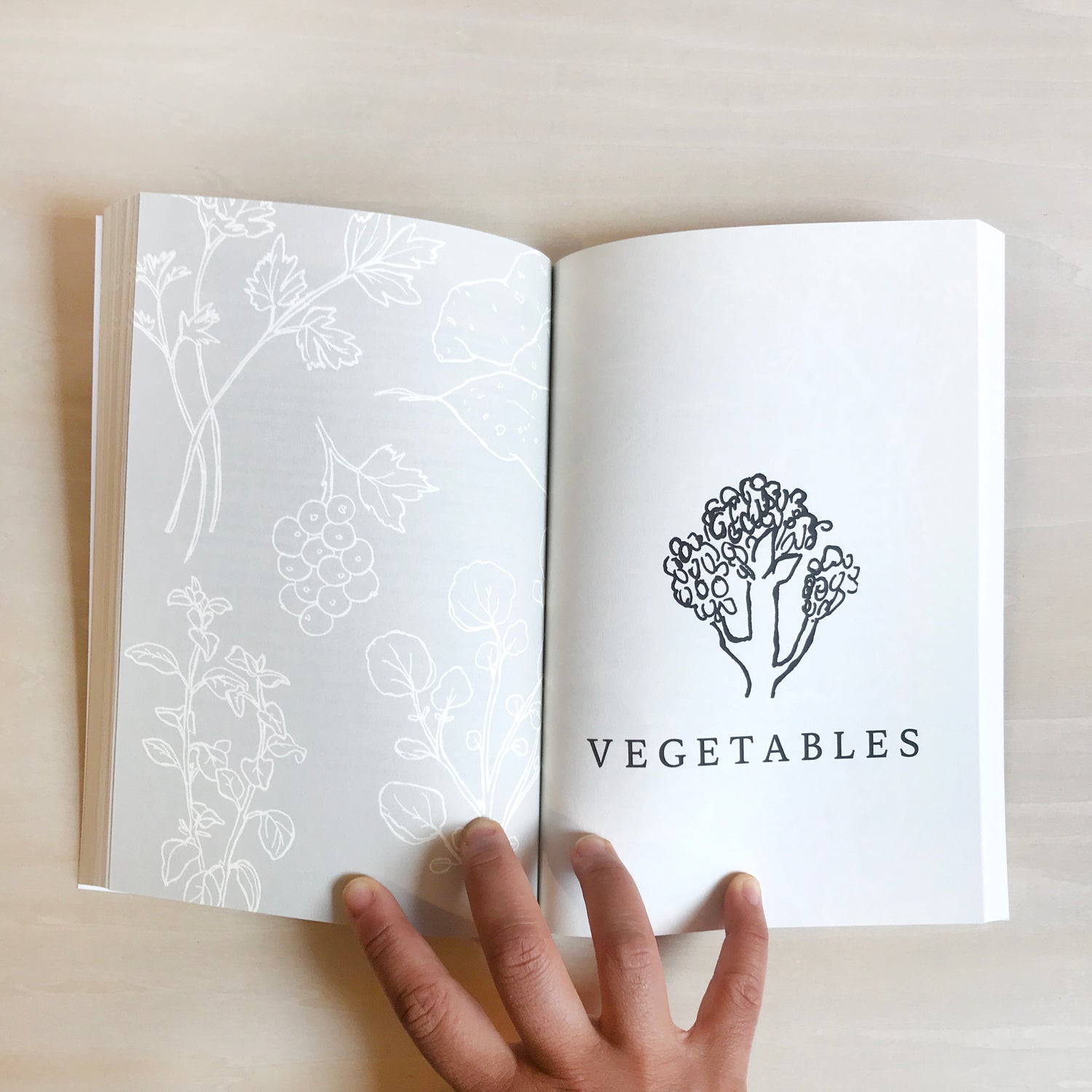"Growing Perennial Foods" book open to "vegetable" chapter page featuring an illustrated broccoli with title below on the right page, light gray and white patterning of herbs on left page with a hand holding the spread open on light wood desk
