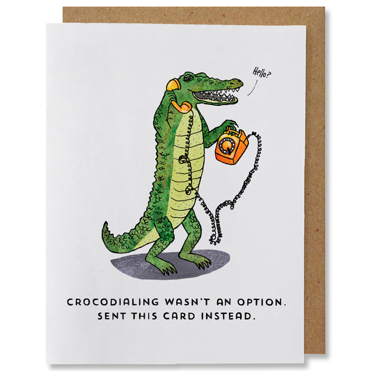 Illustrated everyday greeting card featuring a green crocodile holding an orange rotary phone with the receiver at its ear saying “hello”. The caption reads “crocodialing wasn’t an option. Sent this card instead.”